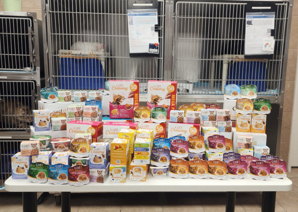 A large collection of Catit Creamy and Catit Dinner cat treat packaging arranged on a table in front of animal cages