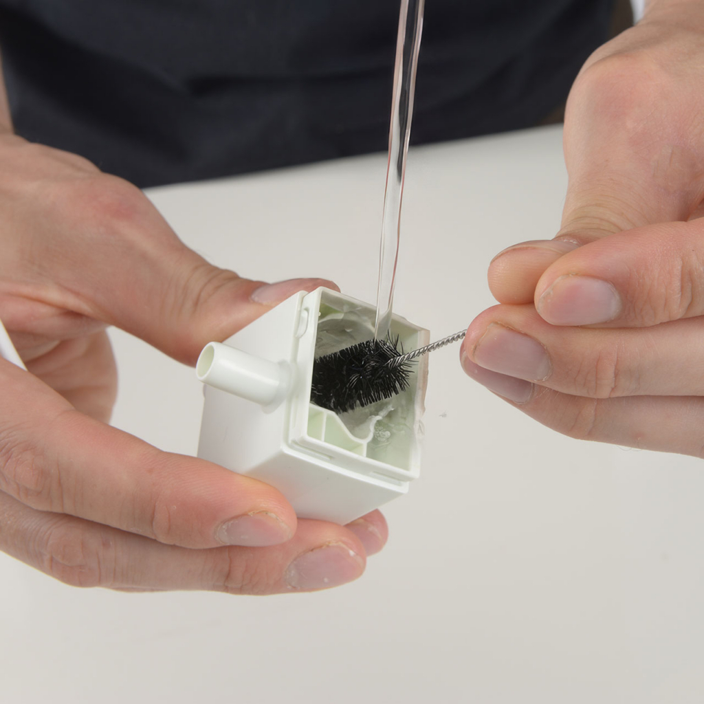 Hands cleaning the interior of a PIXI fountain pump housing with a small brush under running water