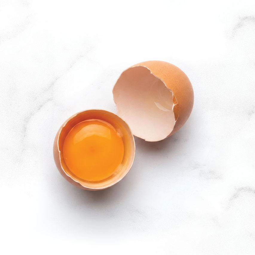 A raw egg cracked open on a white marble surface with the yolk visible in one half of the shell