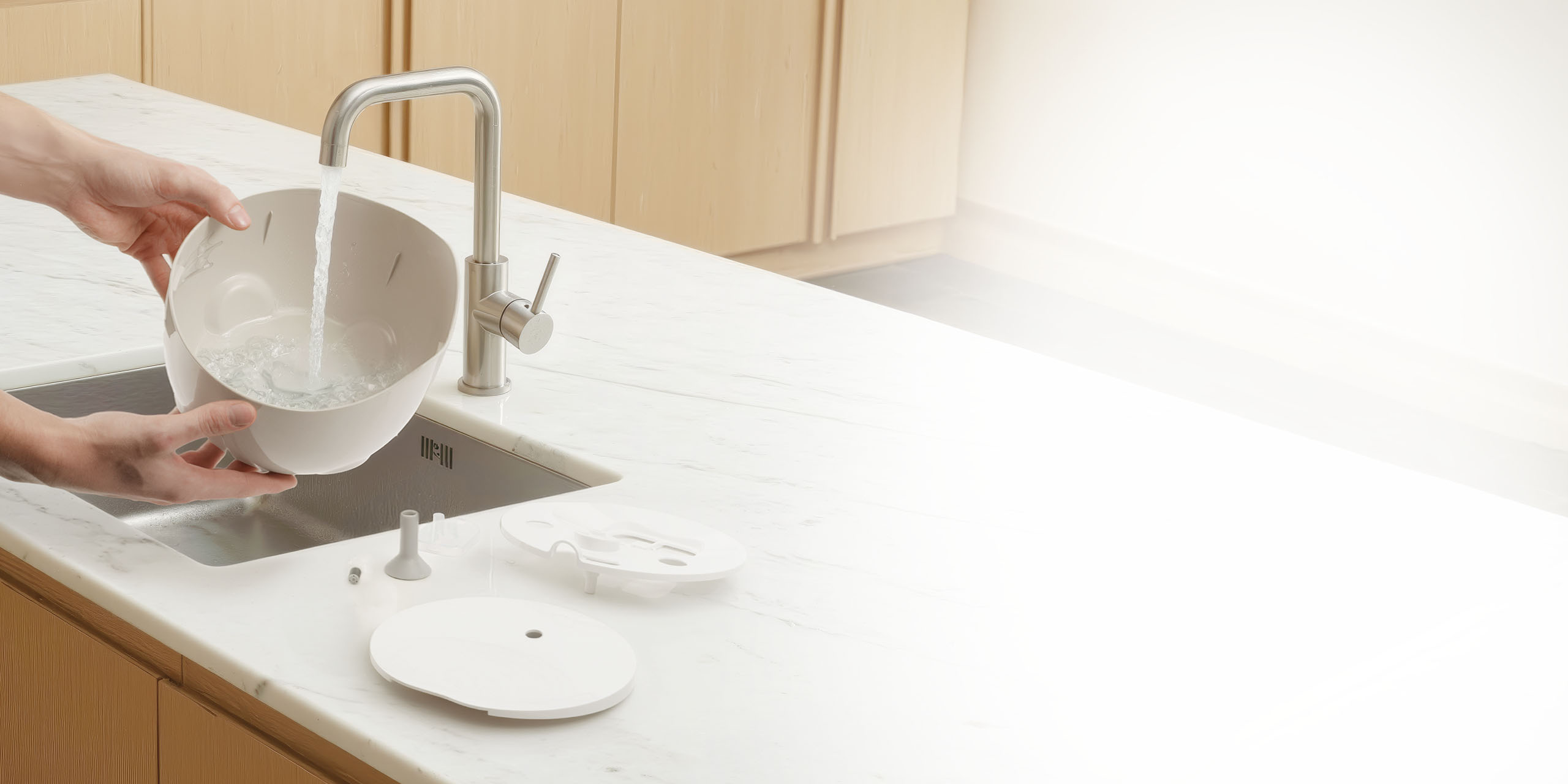 Hands washing the bowl of a Catit PIXI fountain under a kitchen faucet with disassembled parts on the counter