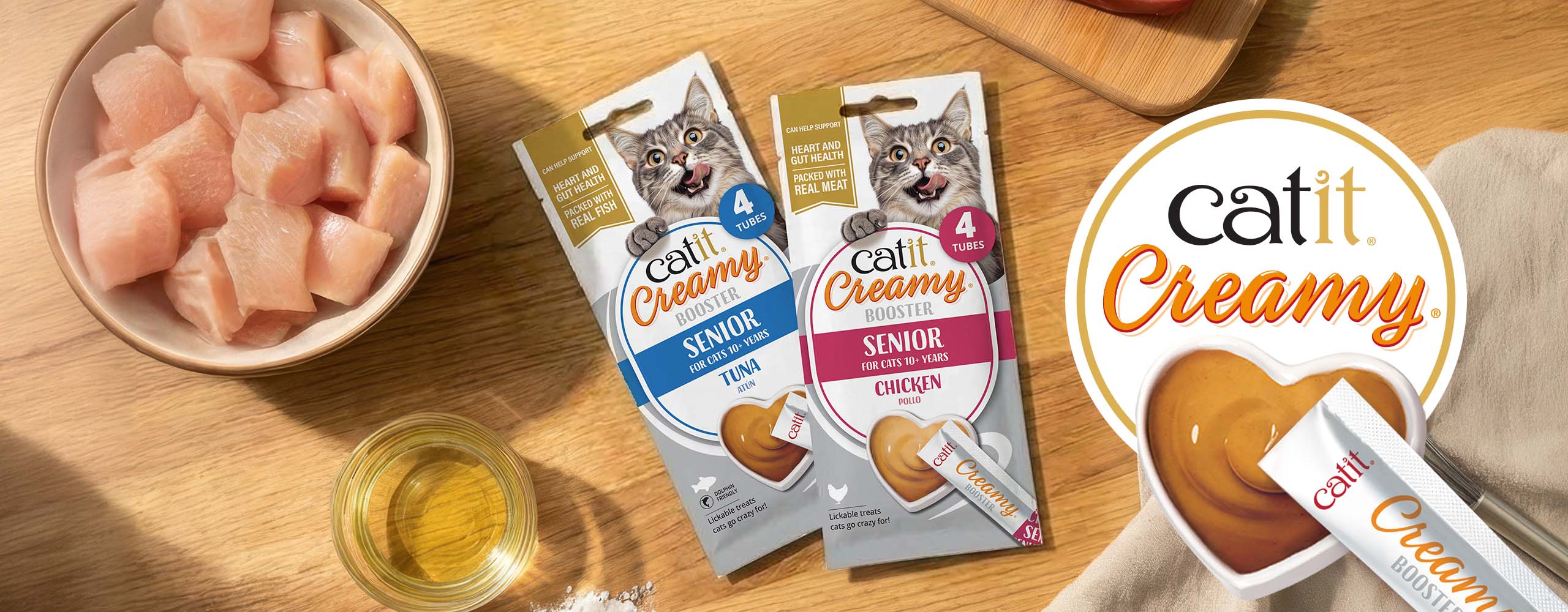 Catit Creamy Booster Senior cat treat packaging in tuna and chicken flavors with a bowl of raw chicken and a heart-shaped dish