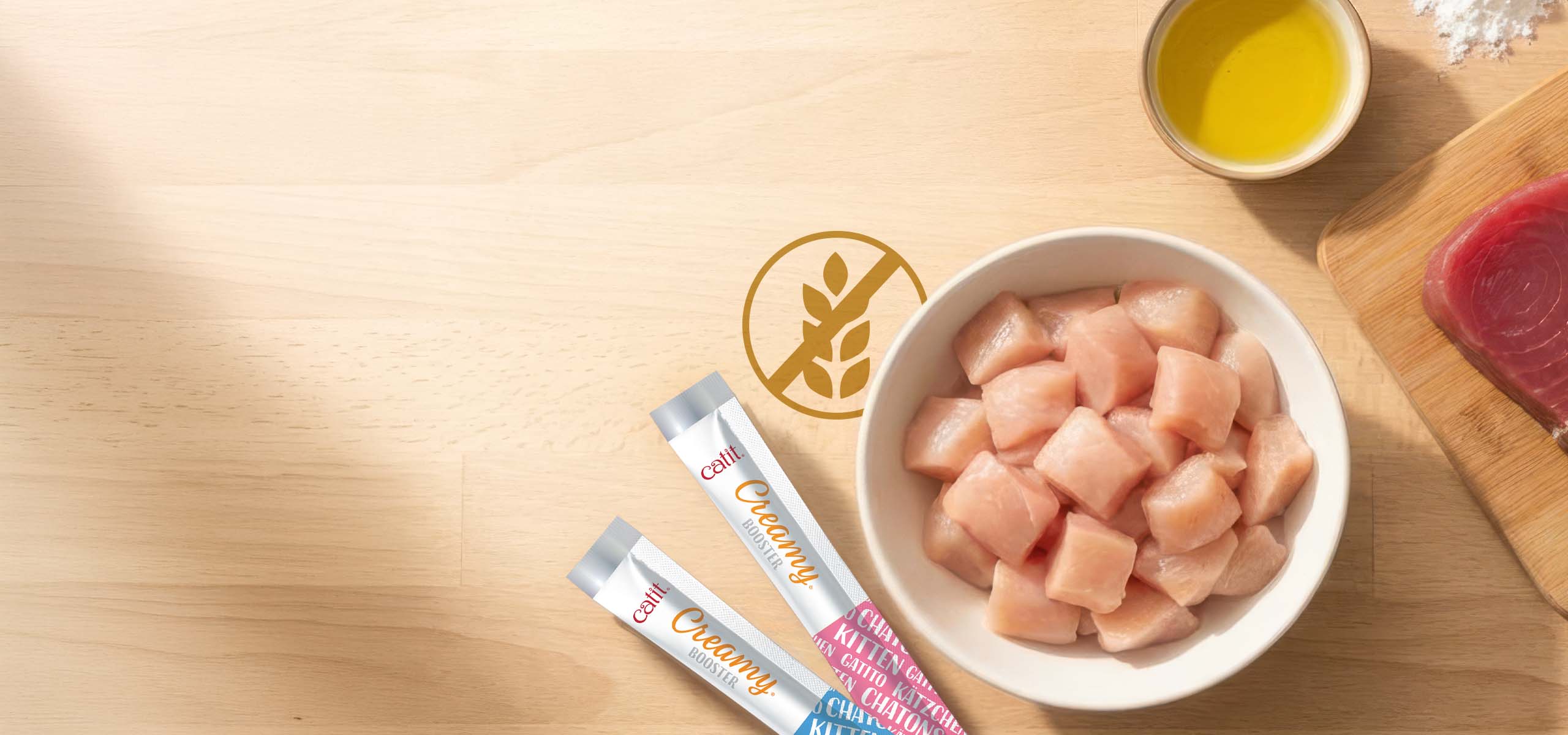 Two Catit Creamy Booster treat tubes next to a bowl of raw chicken cubes and a small bowl of oil