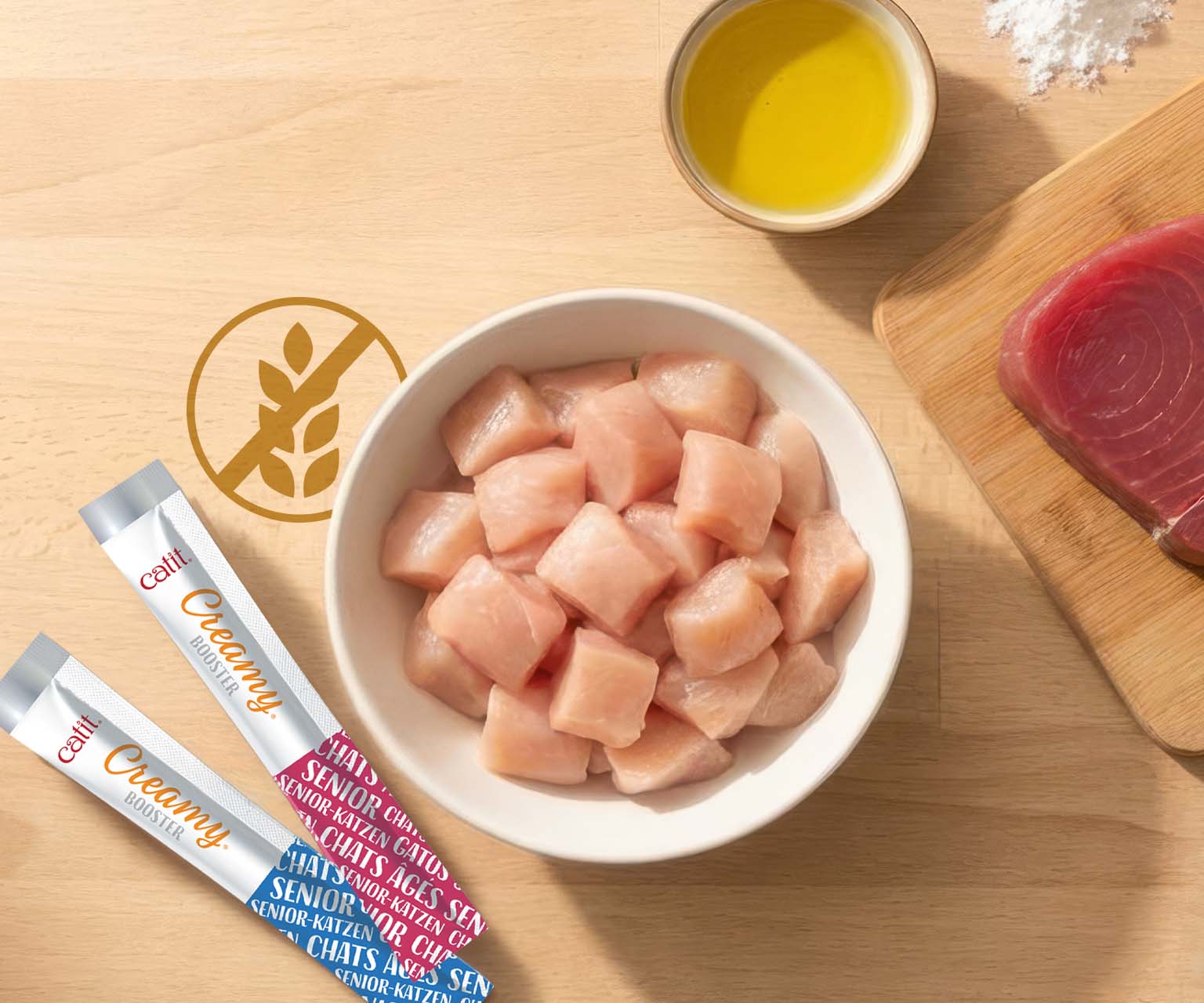 Two Catit Creamy Booster tubes for senior cats next to a bowl of raw chicken cubes, a piece of tuna, and oil