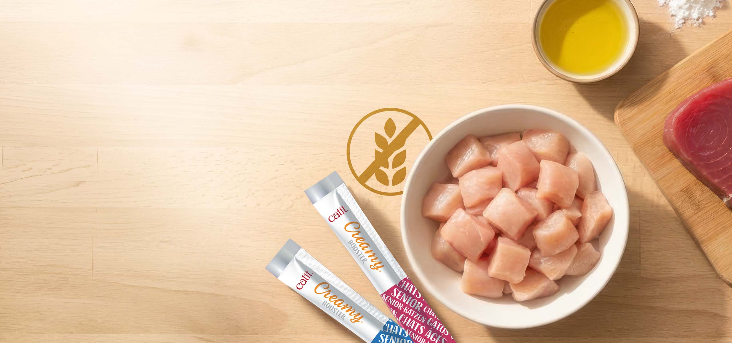 Catit Creamy Booster cat treat tubes next to a bowl of raw chicken cubes and a small bowl of oil