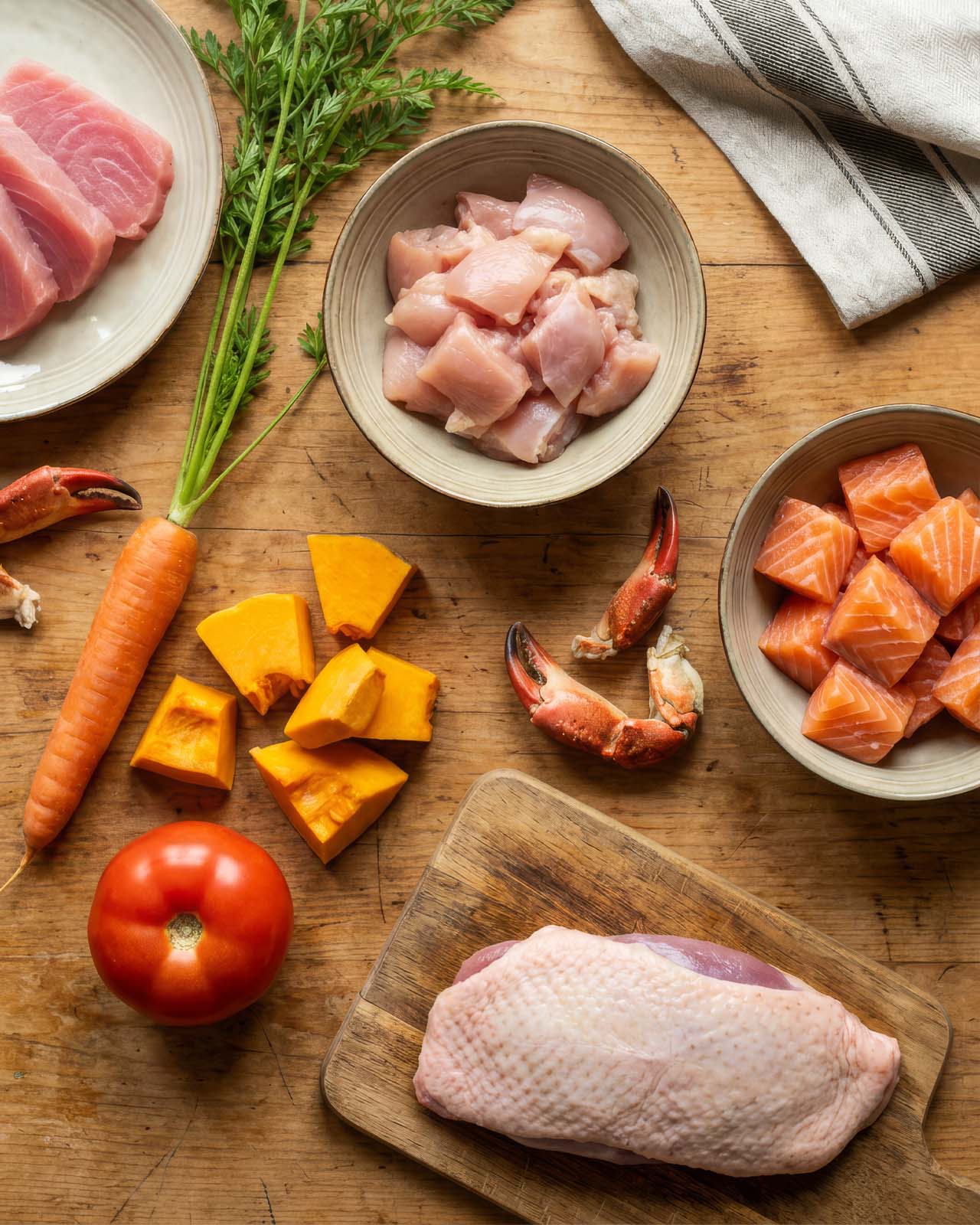 Raw ingredients for cat food including tuna, chicken, salmon, crab claws, carrot, pumpkin, tomato, and a duck breast