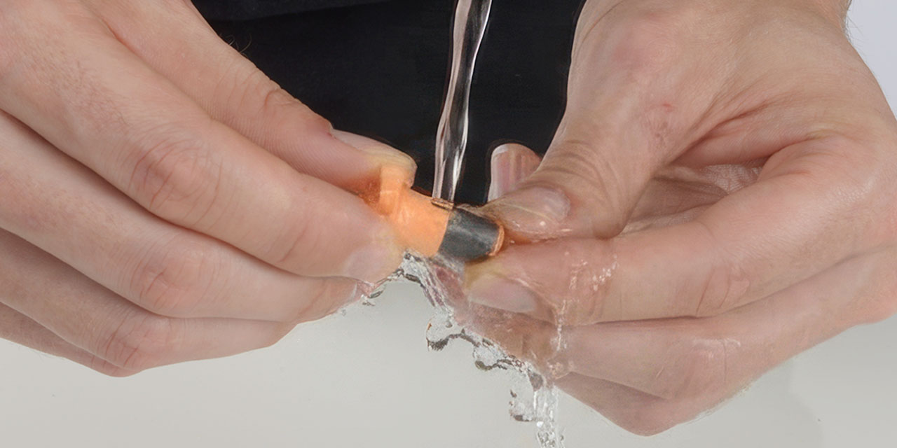 Hands rinsing a small orange and grey plastic part of a Catit fountain under running water