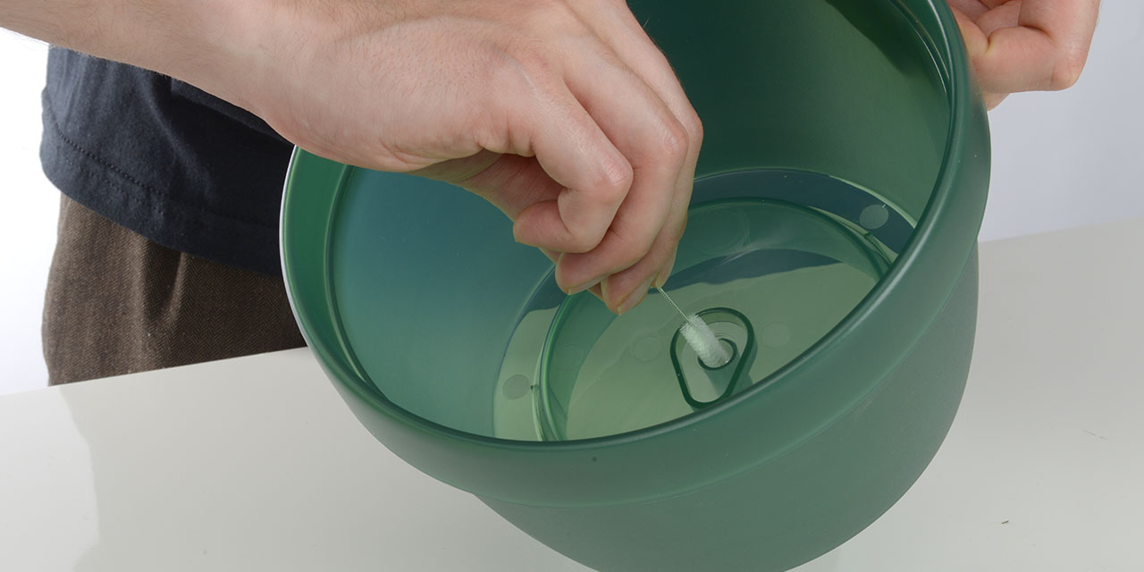 A person uses a small brush to clean the central tube of a green Catit PIXI fountain bowl