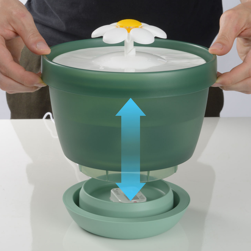A person holding the upper reservoir of a green Catit flower water fountain above its base unit with a bleu arrow