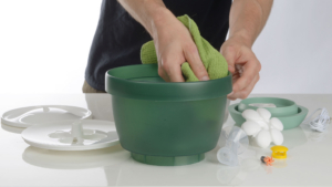 A person cleaning the green basin of a Catit flower fountain with a green cloth, with disassembled parts laid out nearby