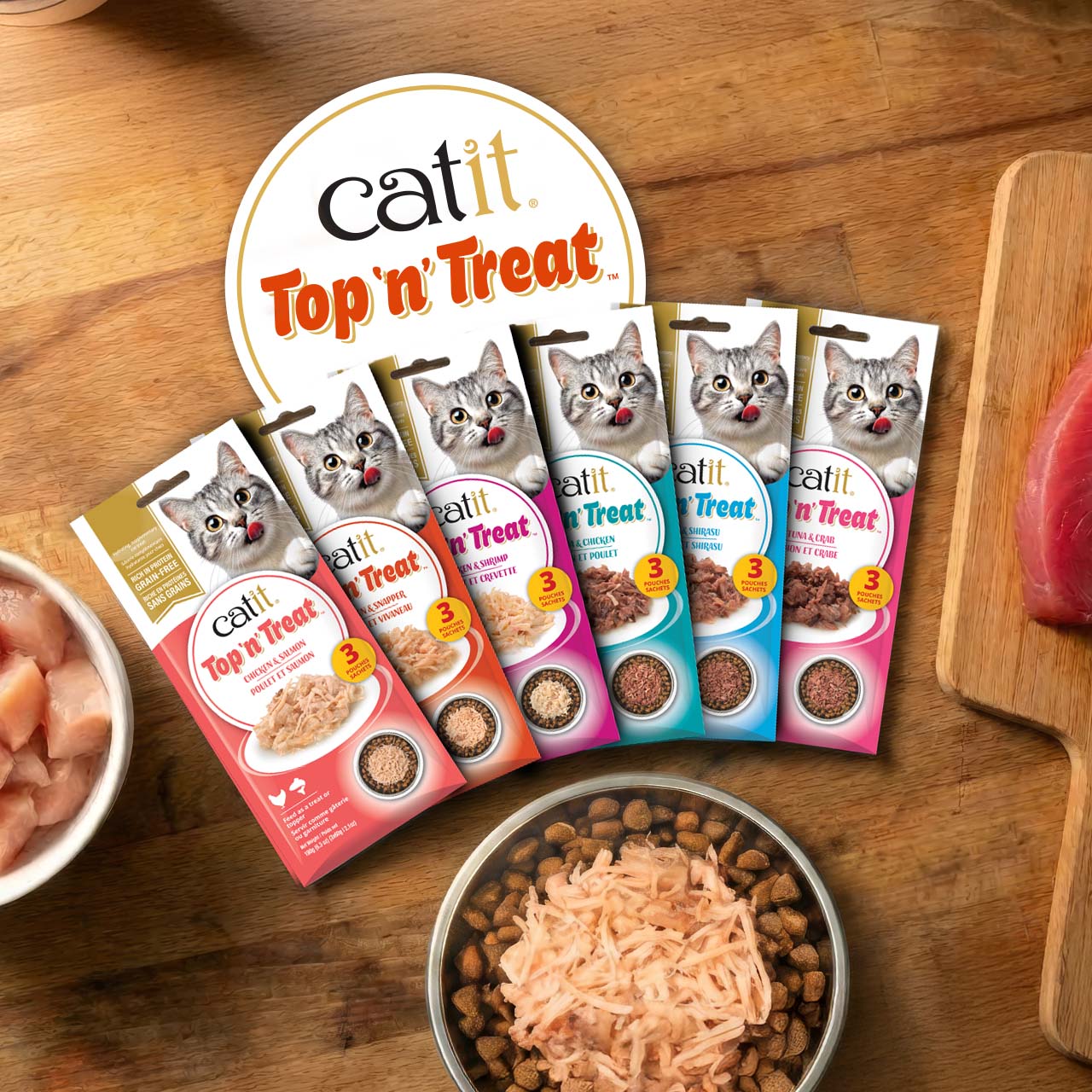 Six packages of Catit Top 'n Treat cat food toppers arranged in a fan shape on a wooden surface with a bowl of dry food
