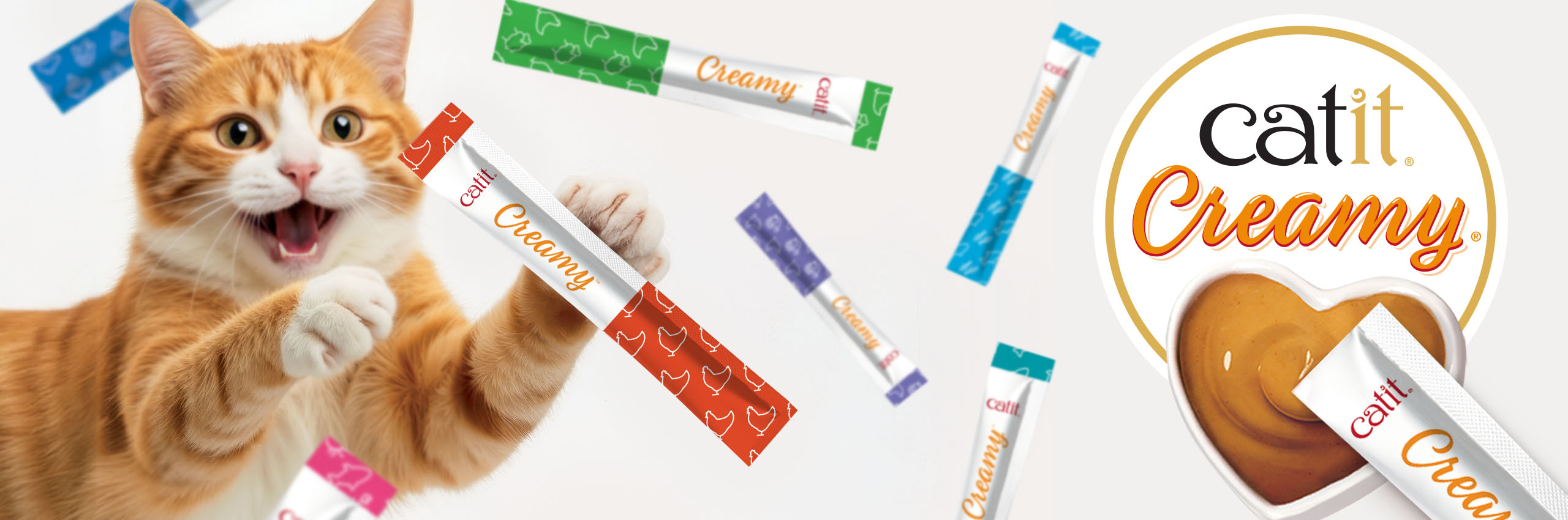 An orange tabby cat excitedly reaching for a Catit Creamy treat tube, surrounded by various flavors of the treat packaging