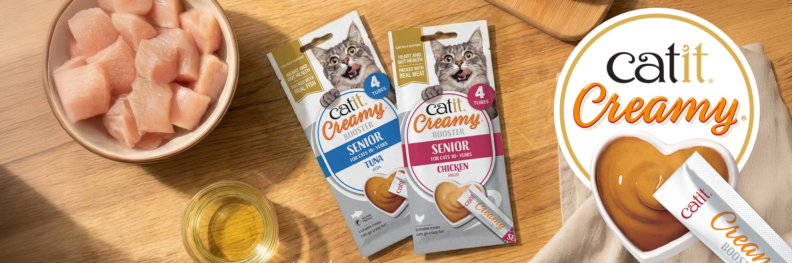 Catit Creamy Booster lickable treats for senior cats in tuna and chicken flavour laying on wooden countertop with ingredients