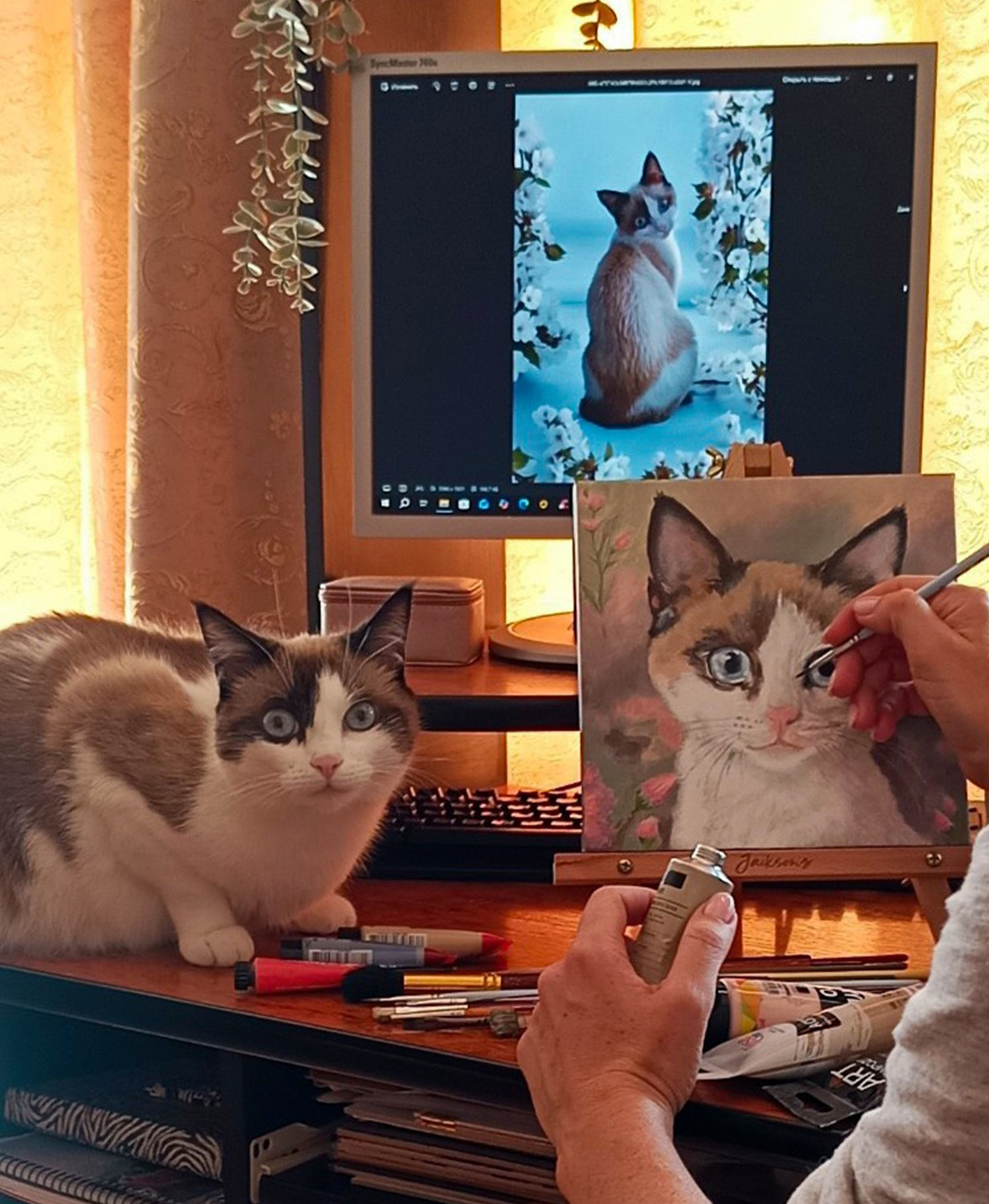 Simfonietta, eine weiß-braune Katze, blickt in die Kamera und sitzt auf einem Schreibtisch vor einem Computerbildschirm, auf dem ihr Porträt zu sehen ist, während ihr Mensch ein Porträt von ihr malt