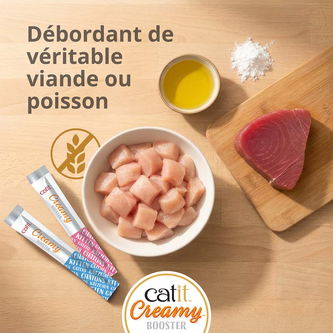 Bol de morceaux de poulet et de thon sur une planche déposée sur un comptoir en bois, avec deux tubes de gâterie Catit Creamy Booster pour chatons