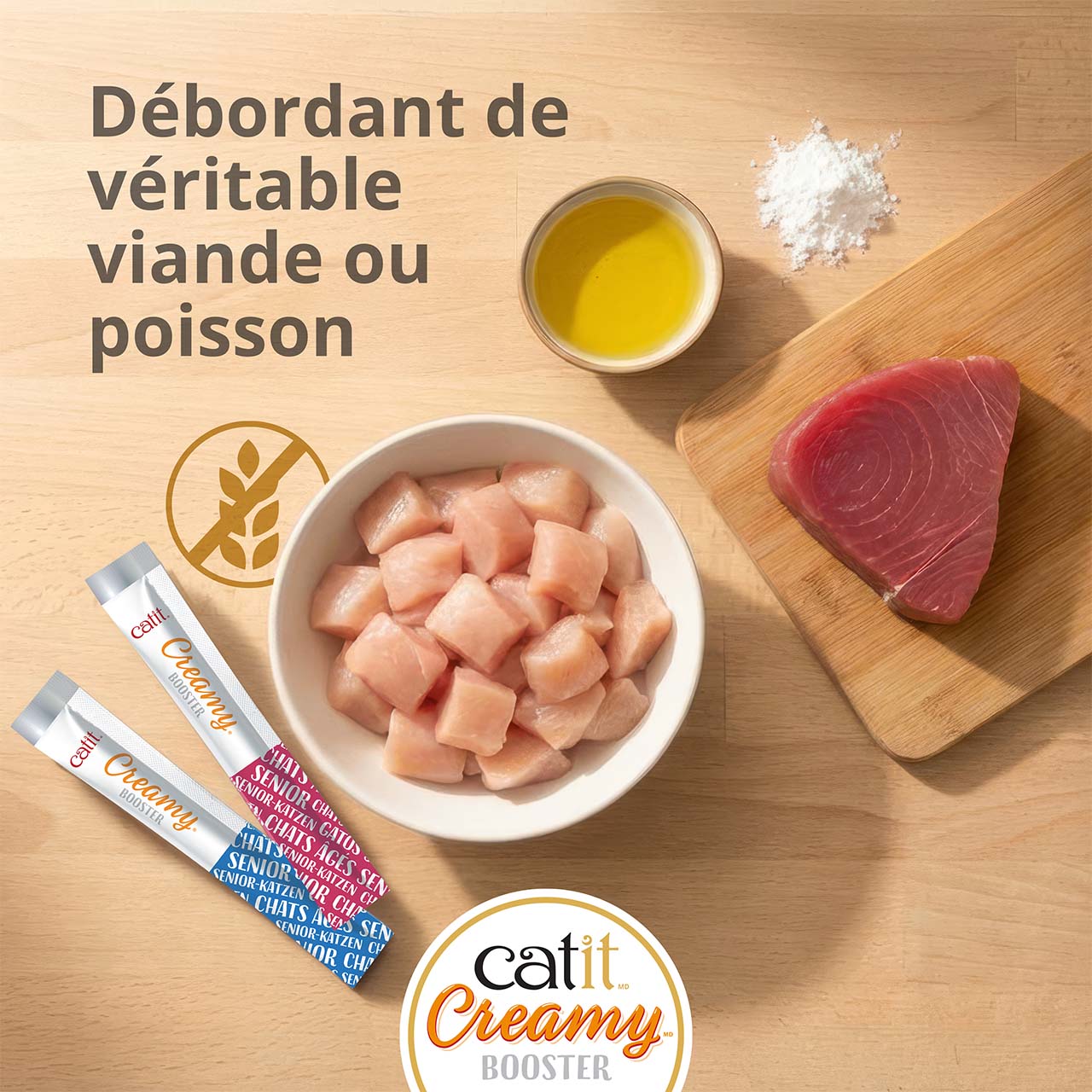 Bol de morceaux de poulet et de thon sur une planche déposée sur un comptoir en bois, avec deux tubes de gâterie Catit Creamy Booster pour chatons
