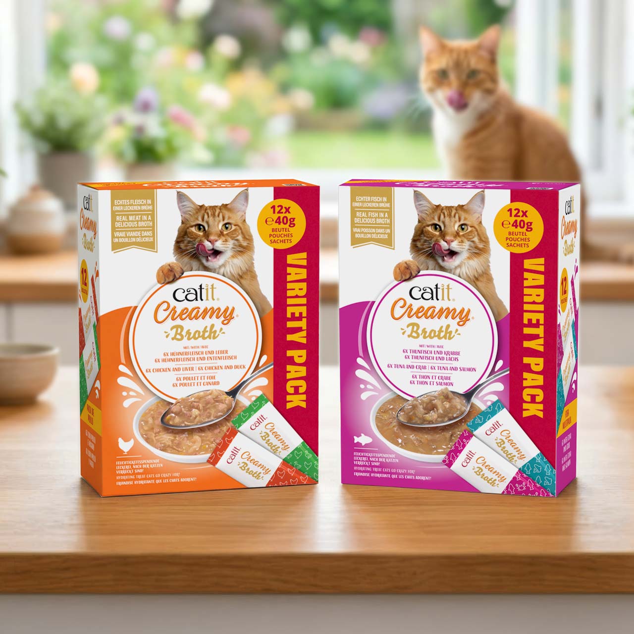 Catit Creamy Broth Sortenmix-Packungen auf einer Küchenarbeitsplatte aus Holz; im Hintergrund blickt eine orangefarbene Katze auf die Verpackungen